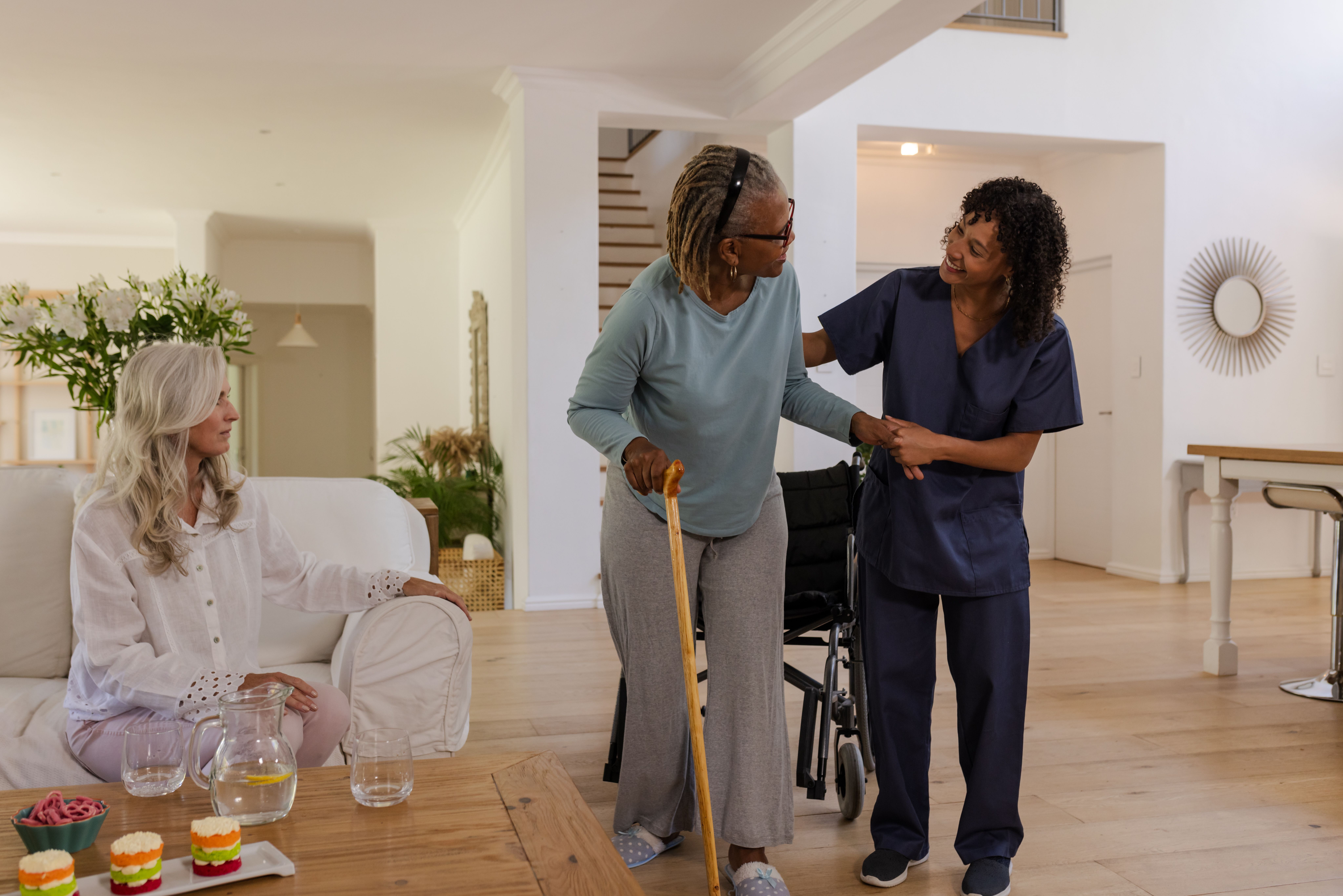 Caregiver assisting senior woman with cane while friend watches in living room. Caregiving, assistance, elderly, companionship, support, healthcare
